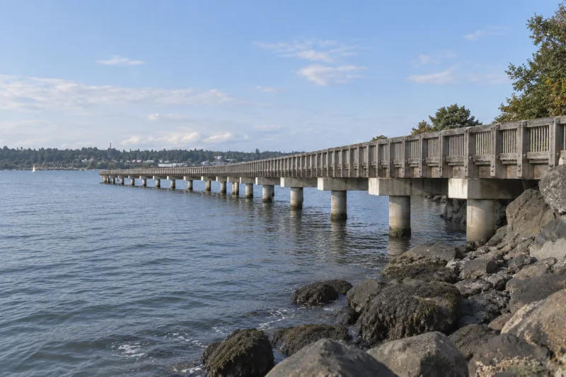The Taylor Dock boardwalk at Fairhaven in Bellingham seen from the shore rocks beside calm water.