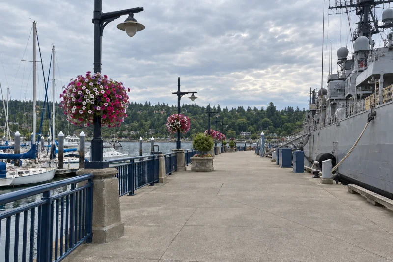 A wide paved boardwalk with blue railings and flower-basket lamp posts beside a grey naval ship and a marina at Bremerton.