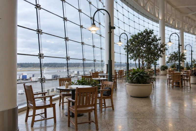 Round tables and wooden chairs beneath the tall curved glass wall of a seating hall at Seattle-Tacoma airport.