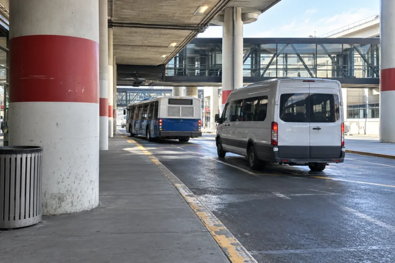 A shuttle van and a blue bus along the terminal kerb under the concrete canopy.