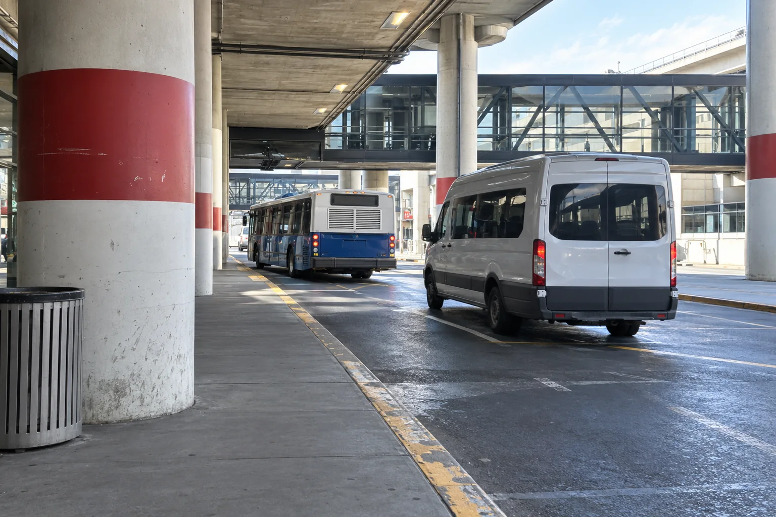 A shuttle van and a blue bus along the terminal kerb under the concrete canopy.
