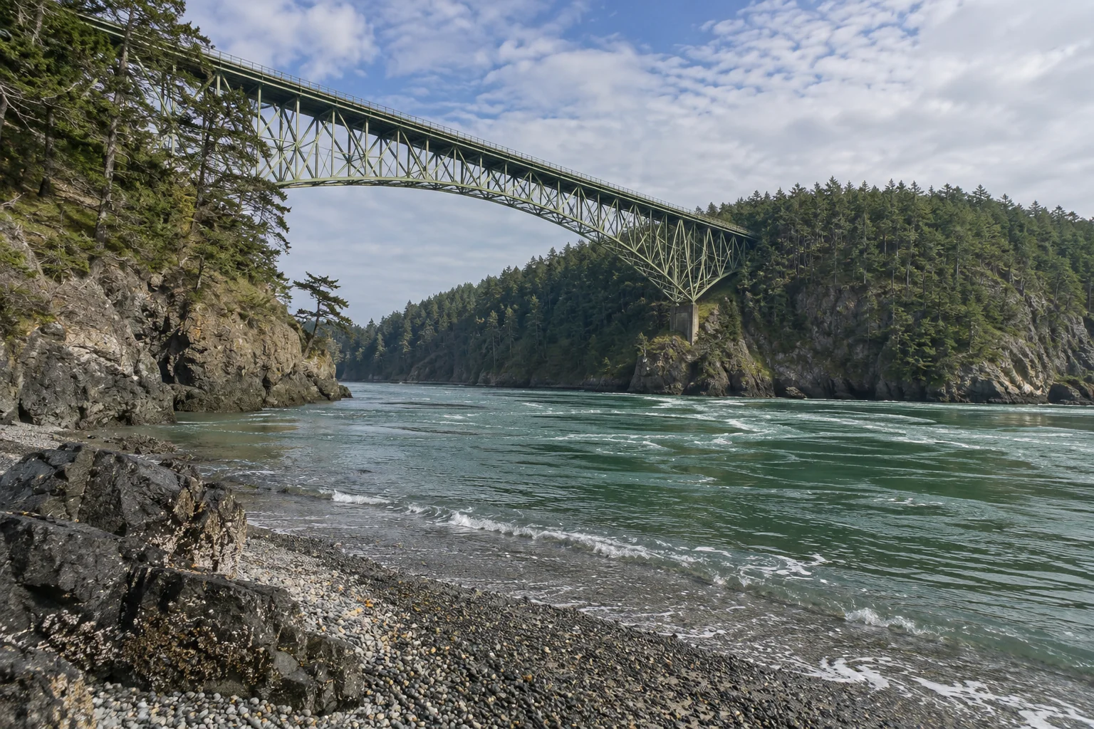 The Deception Pass Bridge, a green steel arch high over the strait, seen from the rocky shore.