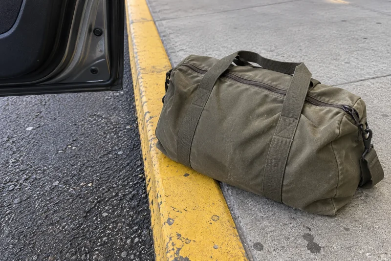 A duffel bag on the departures curb beside the open rear door of a parked car at Seattle-Tacoma airport.