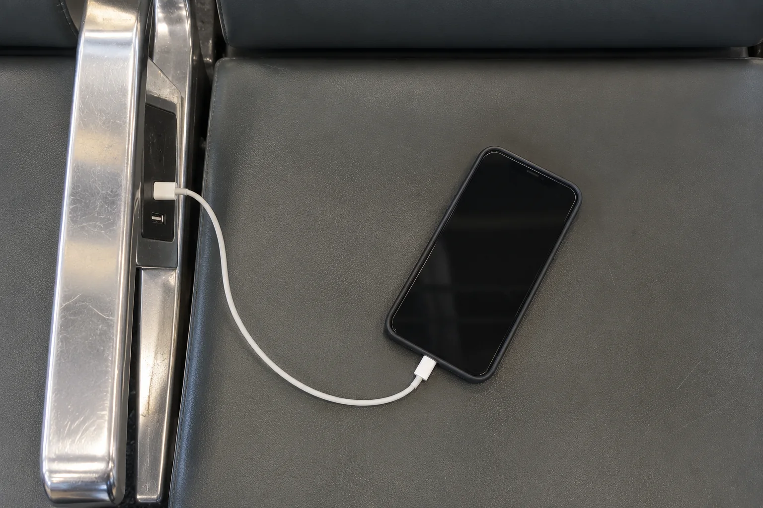 A phone charging from the power outlet on the armrest of a black gate seat at Seattle-Tacoma airport.