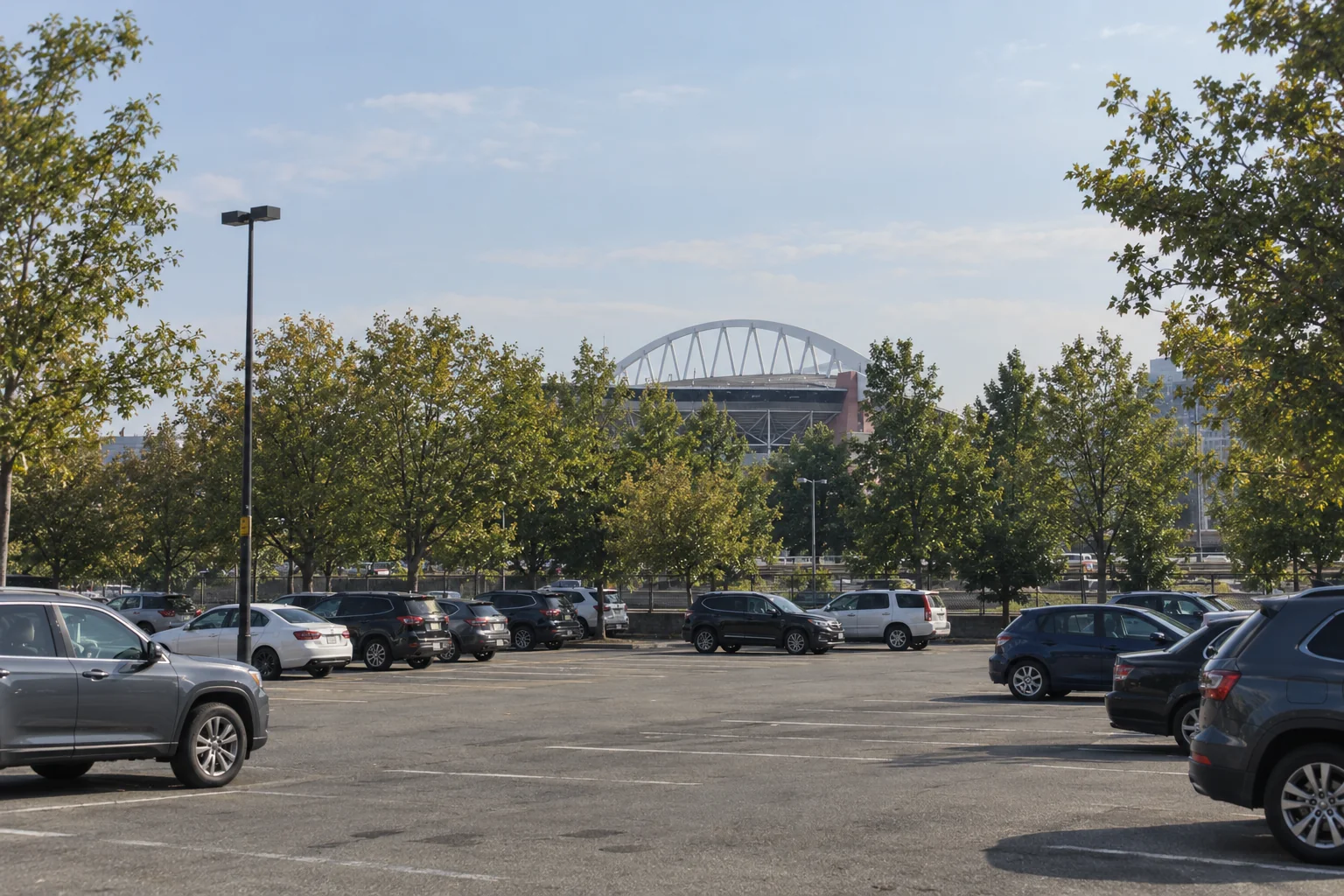 The white steel arch and curved roof of Lumen Field in Seattle seen through street trees across a parking lot.