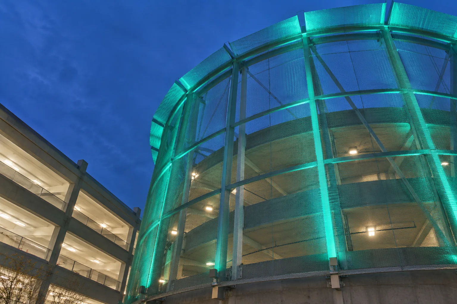 A round ramp drum of the SEA Rental Car Facility glowing teal at dusk, seen from its base