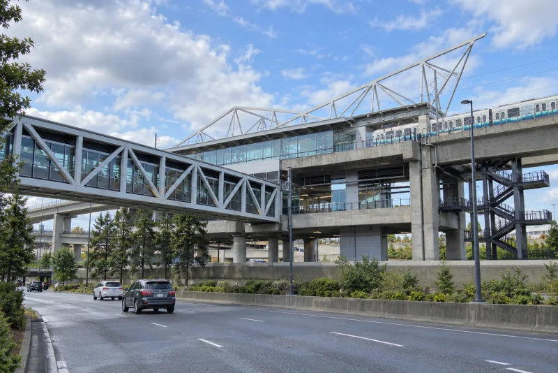 The elevated concrete and glass SeaTac/Airport Link station with its white steel truss canopy and the pedestrian bridge leading to it.