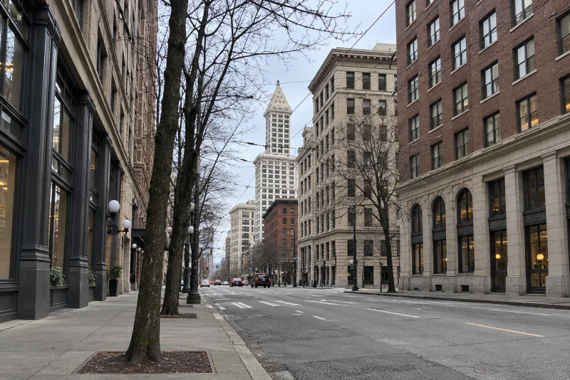A downtown Seattle avenue between tall masonry buildings with overhead trolley wires and a cream tower ahead.